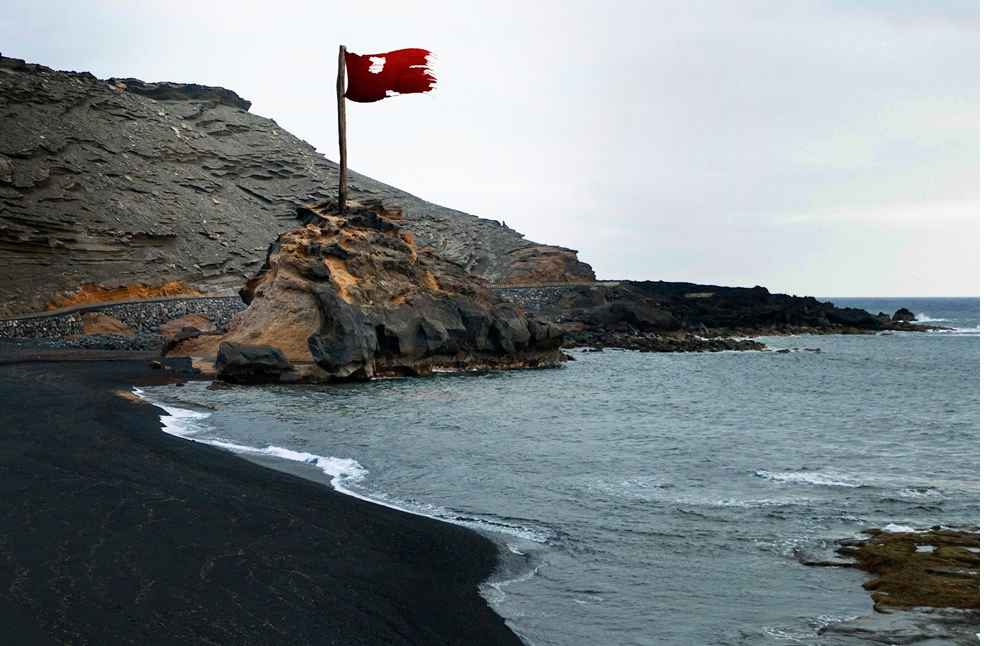 Maritime Crisis 2026: Shipping Losses and Rising Risks 1 Maritime Crisis 2026 represented by a red warning flag flying above a rocky coastline and dark-sand beach.
